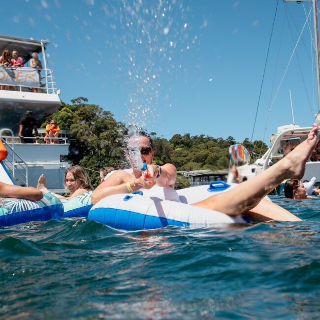 People are enjoying a sunny day on the water, floating on inflatable tubes, with boats in the background. One person is using a water gun, splashing water at what looks like a captivating Catamaran party in Sydney.