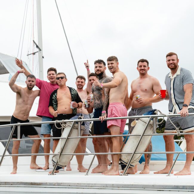 A group of nine men are standing together on the deck of a boat, smiling and posing for the camera. Some are shirtless, while others are in casual summer attire. They appear to be having a lively time, with a few holding drinks. The background shows part of the boat and lush trees.