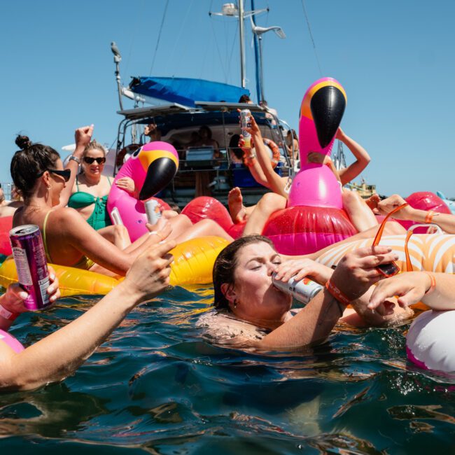 People on inflatable pool floats enjoy drinks in the water near a catamaran party Sydney on a sunny day.