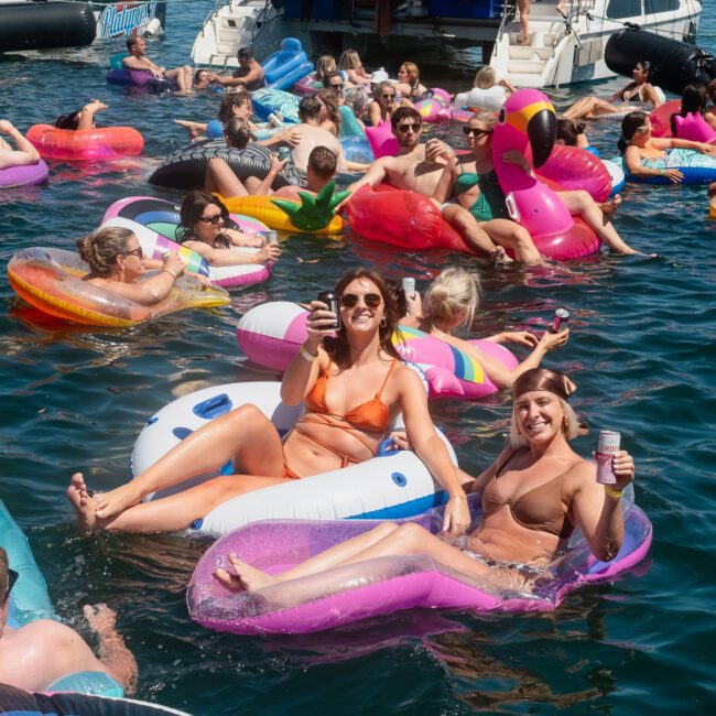 A group of people in swimsuits enjoying a sunny day on various inflatable floats in the water, with luxury yachts and boats in the background.