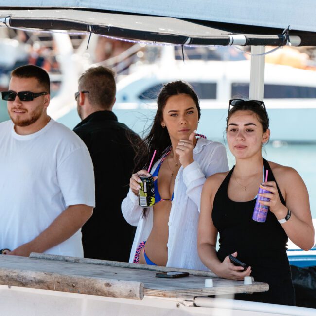 A group of people stands on a boat under a canopy. Two women in swimwear are holding drinks with straws, one in a black top and the other in a white shirt over her swimsuit. A man in a white t-shirt, sunglasses, and flip flops also holds a drink. Other boats are in the background.