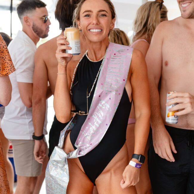 A smiling woman in a black swimsuit wearing a sash that reads "Birthday" holds a drink can. She is surrounded by people in swimwear, creating a festive atmosphere on the yacht under the bright sun.