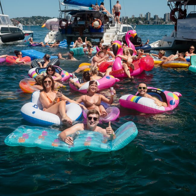 A group of people floating on colorful inflatables in the water, surrounded by boats, and enjoying a sunny day, with a lively DJ boat hire Sydney adding to the fun atmosphere.