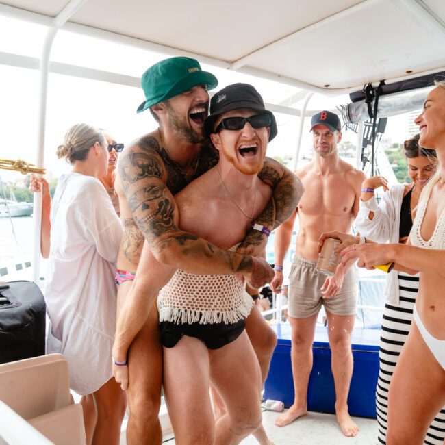 A group of joyful people are on a boat having fun. One person with tattoos is hugging another from behind, both wearing hats and smiling. Others in the group are chatting and laughing, capturing selfies to remember the moment. They are dressed in beachwear, and the background shows a marina with boats.