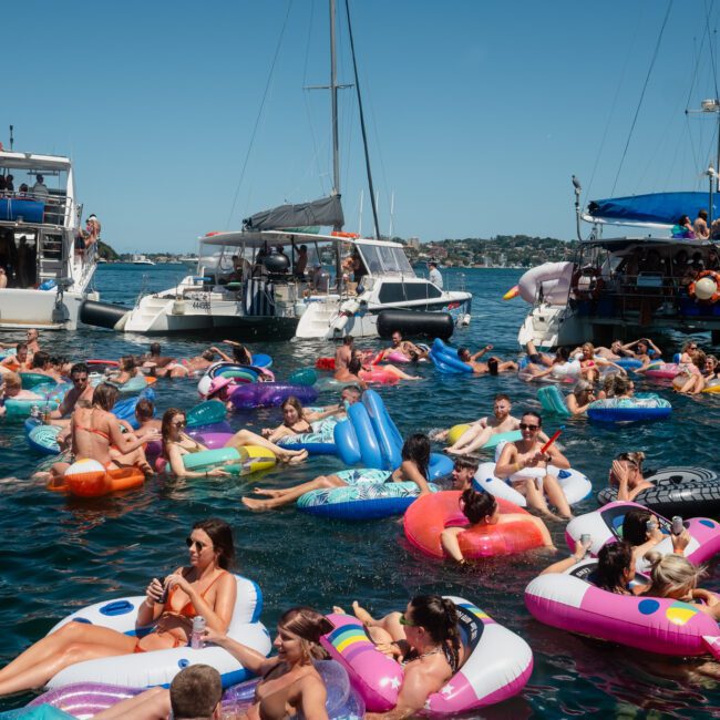 A group of people floats on inflatable rafts and toys on a body of water near several docked boats on a sunny day, creating the perfect backdrop for a Sydney boat party hire.
