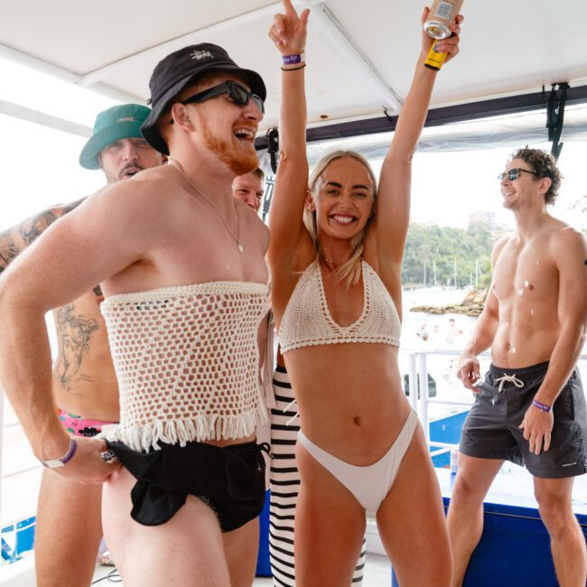 A group of people on a boat enjoys a lively party. Two individuals in the foreground wear crocheted tops and swimwear, with the woman raising her hands and smiling radiantly. Others are casually dressed, holding drinks, and mingling. Pristine trees and water set a beautiful backdrop.