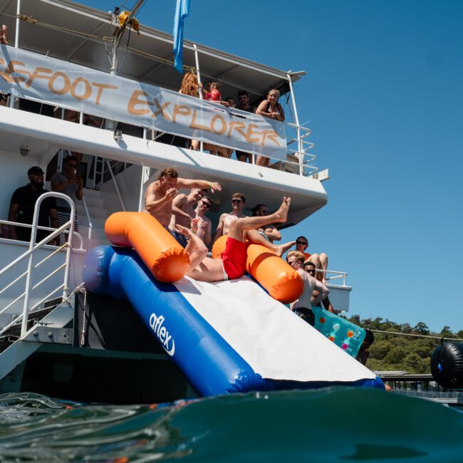 People are sliding down an inflatable slide from a boat named "Barefoot Explorer" into the water, with others watching from the boat. Enjoy the fun and excitement of a Private yacht charter Sydney Harbour near a wooded shoreline under a clear sky.