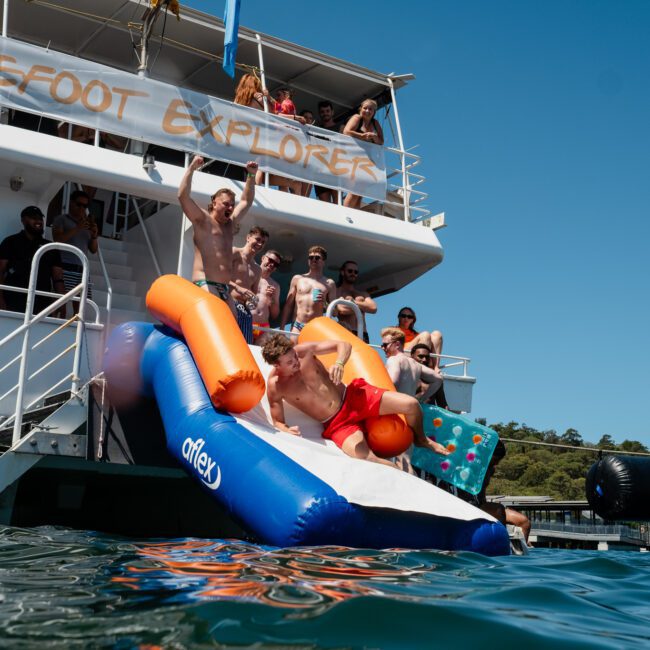 People on a boat watch as one person slides down an inflatable slide into the water, connected to the boat's deck. Clear blue sky and calm water provide a perfect backdrop for this corporate boat event in Sydney.