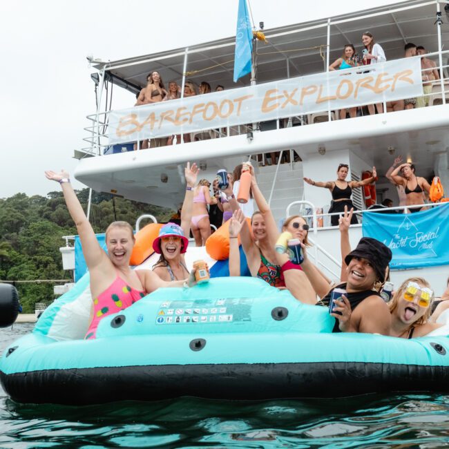 A group of people relax on an inflatable raft in the water, smiling and raising their arms. The raft is in front of a large boat named "Barefoot Explorer," adorned with vibrant flags. More people are visible on the boat, enjoying the scenery with trees lining the shore in the background.