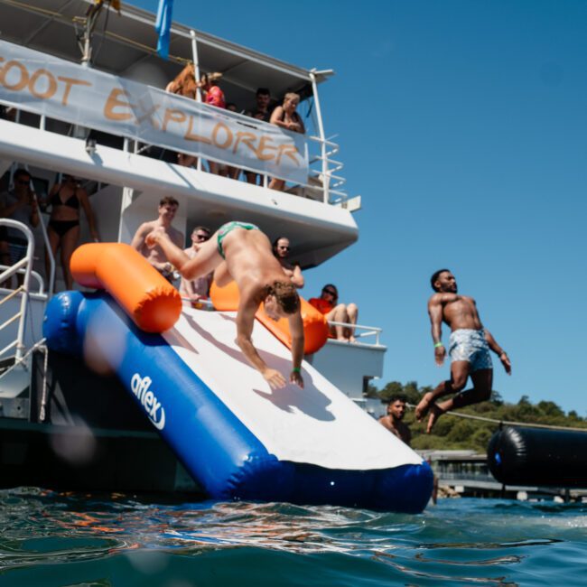 Several people are on the deck of a boat named "Feet Explorer" as some slide off an inflatable slide and others jump into the water, enjoying a corporate boat event in Sydney.