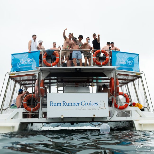 A group of people stand and sit on a two-deck boat named "Rum Runner Cruises," celebrating with raised arms. The boat features life rings, two blue banners, and the tagline "It's all about the Vibe." The calm water enhances the cheerful atmosphere.