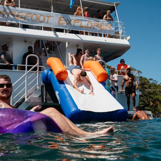 A man slides into the water from a boat with "Barefoot Explorer" written on a banner. Others are using inflatables and socializing on the boat and in the water on a sunny day, enjoying a luxury yacht hire Sydney experience.
