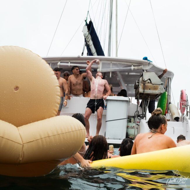 A cheerful boat party scene with many people. Some are on the boat, others are enjoying the water. An inflatable float in the shape of a brown armchair and a yellow duck ring add to the fun. One person in black shorts on the boat is pouring a drink over their face.