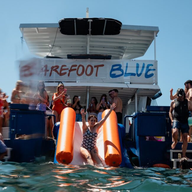 People enjoying a sunny day on a boat named "Barefoot Blue." Some passengers are using the slides to enter the water while others are lounging and socializing on the boat. Experience an unforgettable time with Sydney boat party hire, combining fun and relaxation in one amazing outing.