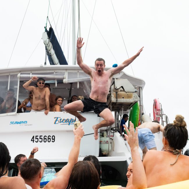 A group of friends are having fun on a boat named "Escape." A man with a surprised expression is jumping off the boat into the water, arms outstretched. Others are on the boat and in the water, cheering and enjoying the moment.