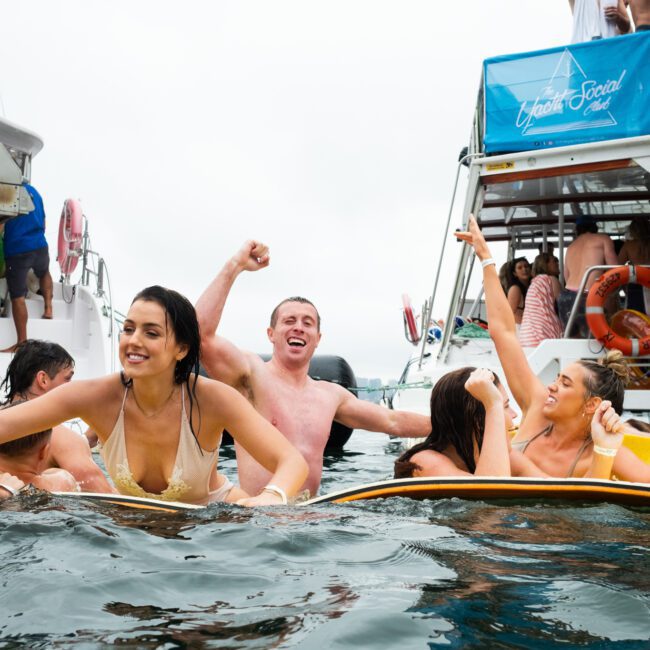 A group of people in swimsuits joyfully hanging out in the water between two boats, celebrating with smiles and raised arms. The setting appears to be a lively social gathering on the water. One boat proudly displays a "Yacht Social Club" banner, enhancing the festive atmosphere.