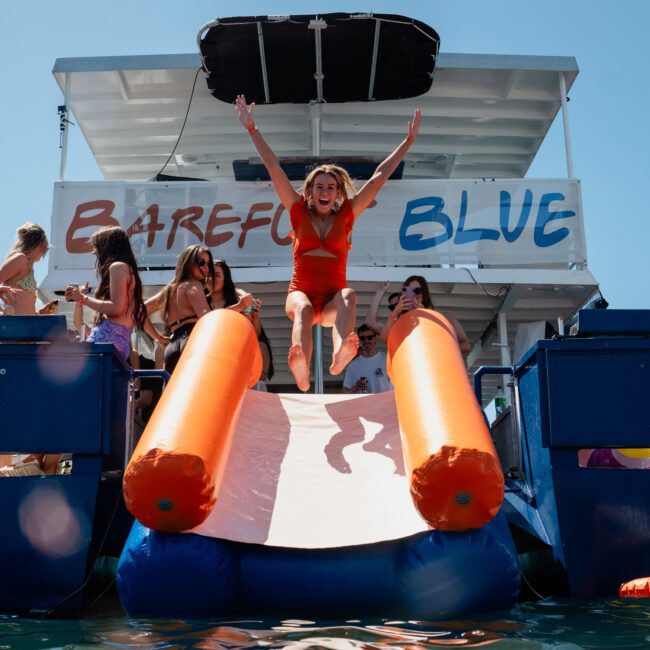 A woman in a red swimsuit is sliding down an inflatable slide into the water from a boat with a "Barefoot Blue" banner. Other people in swimsuits are on the private yacht charter in Sydney Harbour, watching and enjoying the scene.