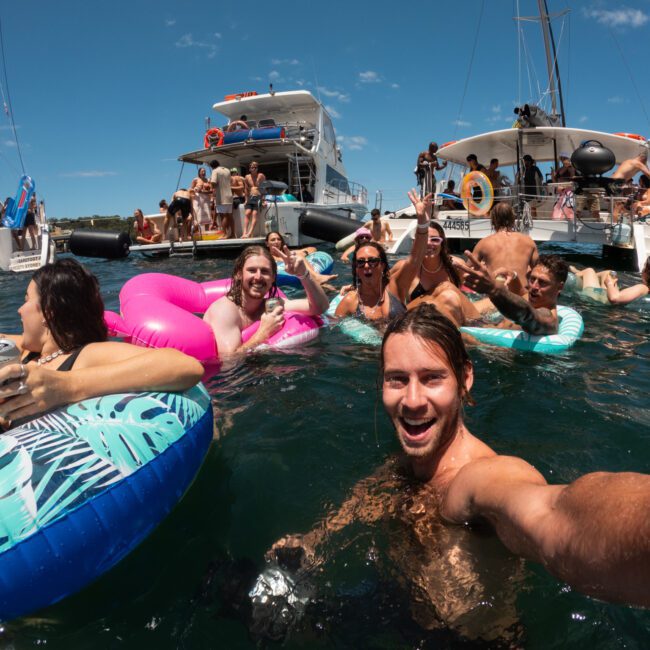 A group of people enjoying a day on the water, lounging on colorful inflatable rings and floats. They are surrounded by boats, with some people waving and smiling at the camera. The sky is clear blue, and everyone appears to be having an amazing time under the warm sunshine.