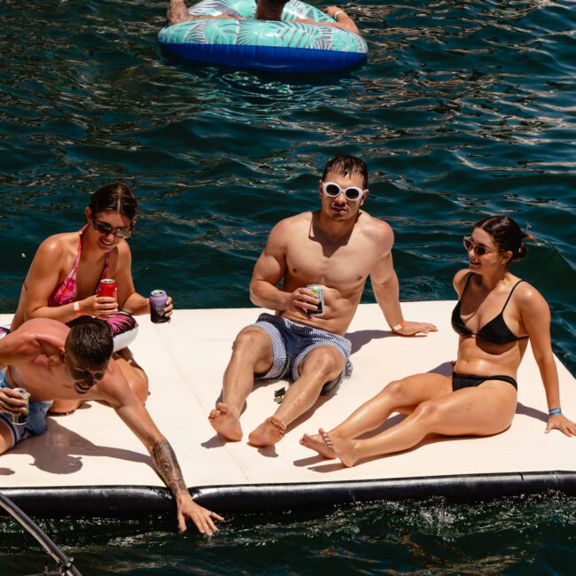 Four friends are lounging on a floating platform in a serene body of water, savoring drinks and the bright, sunny weather. Two men and two women in swimwear are enjoying the moment: one is lying down, another dips their hand into the water, while two sit together, smiling and chatting.