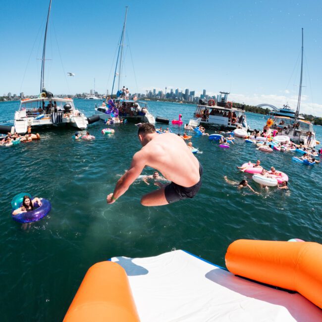 A man in black shorts jumps into a vibrant blue body of water from an orange and white float. Several people, boats, and floaties are visible around him, with a city skyline and clear blue sky in the background. Various inflatables add to the lively atmosphere of this summer getaway.
