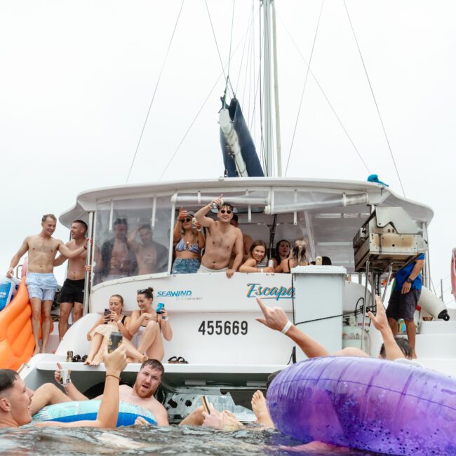 A group of people are enjoying a day on a boat. Some are standing on the deck, dancing and posing for photos, while others are in the water near the boat with inflatables. The boat's name "Escape" is visible, and the atmosphere is lively and joyful.