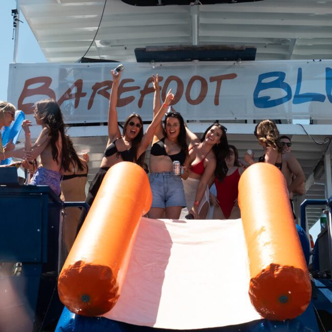 Several people stand and sit on a boat with a slide into the water, enjoying a festive catamaran party in Sydney. Most are wearing swimsuits, smiling and posing, with a sign in the background that reads "BAREFOOT BLU." The scene is vibrant and cheerful.