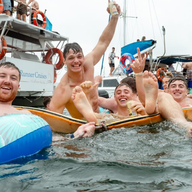A group of four young men are enjoying themselves in the water, holding drinks and laughing. They are surrounded by boats and other people partying. One man is holding his arm up in excitement, while a blue inflatable and other colorful flotation devices bob nearby.