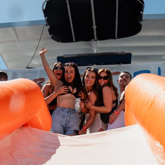 A group of four people, three women and one man, are posing and smiling at the top of an inflatable slide on a sunny day during a Sydney boat party hire.