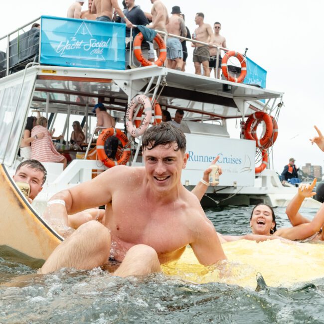 A group of young people enjoys a lively party on a boat labeled "Ghost & Grits" and "Runner Cruises," while others have fun in the water nearby, holding drinks, cheering, and smiling enthusiastically. The atmosphere appears festive and energetic with vibrant laughter echoing across the waves.