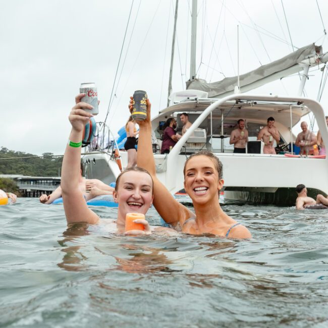 Two people in swimsuits are smiling and holding up drinks while standing waist-deep in the water. Behind them, a boat with several others relaxing onboard adds to the fun vibes. The setting captures a perfect, casual day on the lake, complete with floating devices for extra enjoyment.