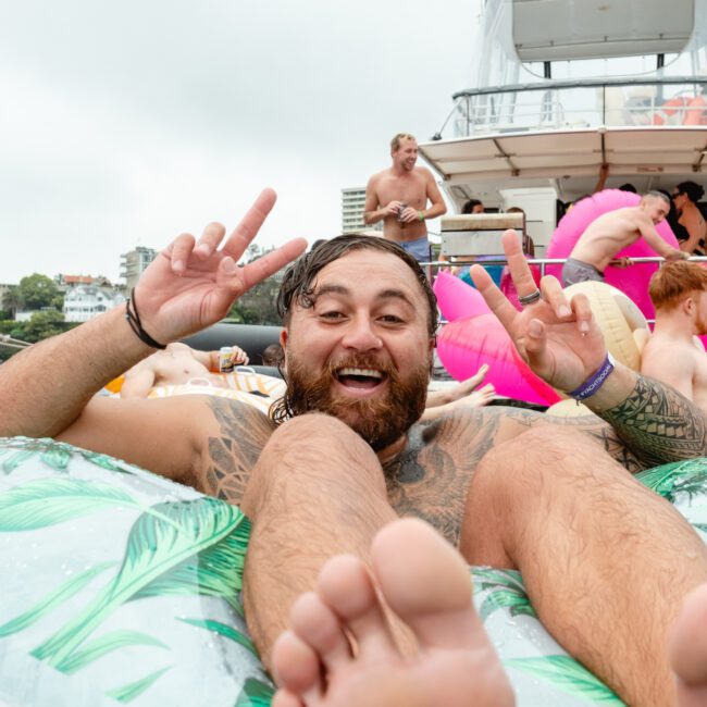 A bearded man with tattoos floats on an inflatable raft, gesturing peace signs with both hands. In the background, people are socializing on a boat adorned with vibrant inflatables, including a pink flamingo. The atmosphere exudes a festive and joyful vibe, enhanced by lively music playing from the boat.