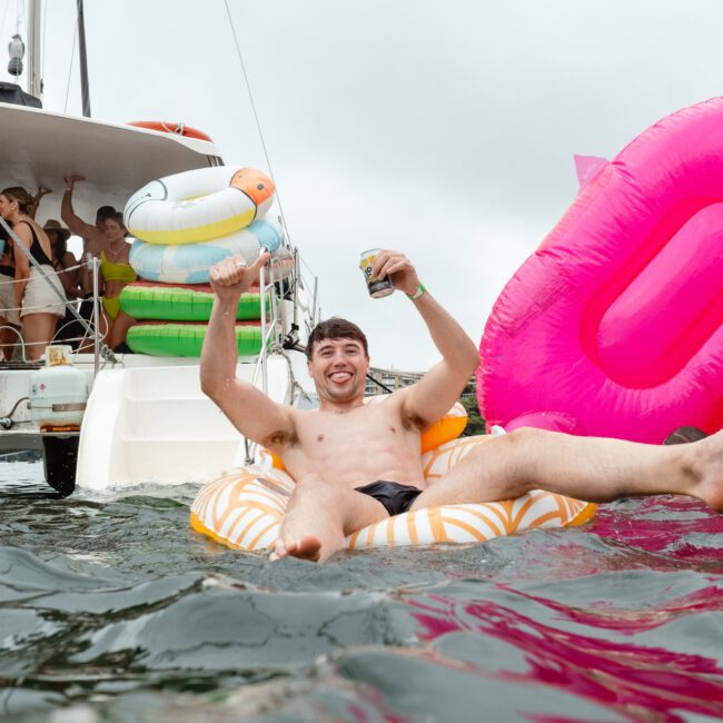 A man relaxes on a float in the water, holding a drink and raising his arm in celebration. Behind him, people on a docked boat enjoy the festivities with vibrant and colorful inflatables all around. The atmosphere is lively and festive, perfect for a joyful summer day.