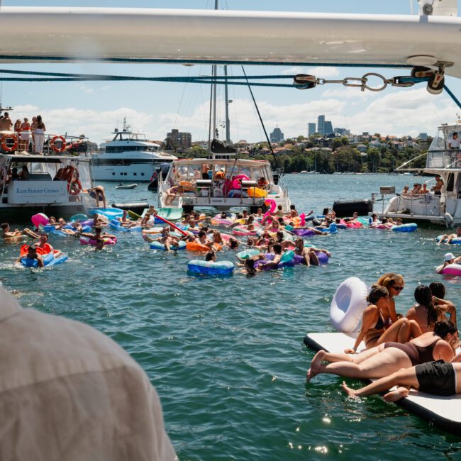 People on inflatables and swimming near yachts in a harbor on a sunny day, enjoying the vibrant atmosphere of a Sydney boat party hire, with some lounging on floating platforms.