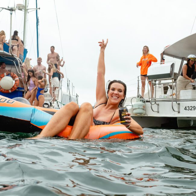 A woman floats on an inflatable tube in the water, holding a drink and raising one arm joyfully. In the background, people are gathered on a vibrant boat, some preparing to jump into the water. The scene embodies a lively, social atmosphere.