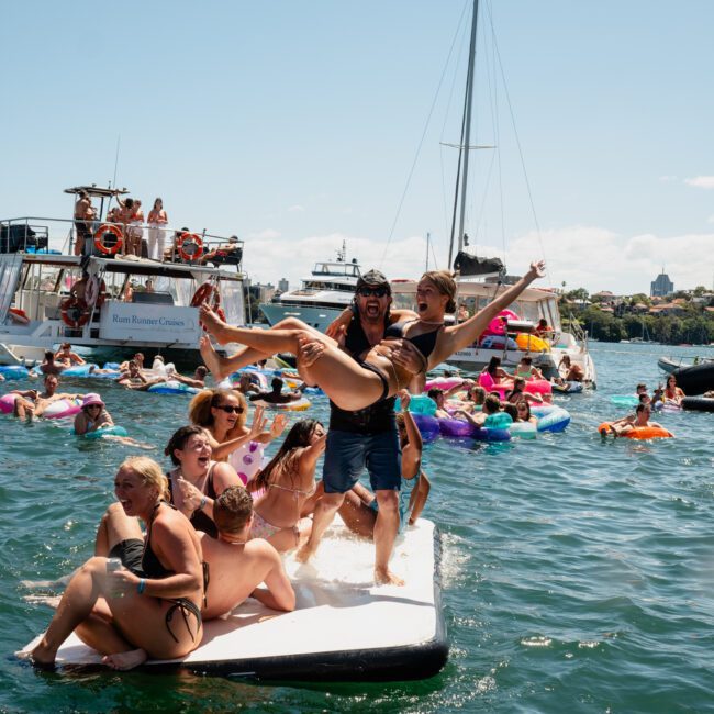 A group of people in swimwear having fun on floats and inflatables in a body of water on a sunny day; a woman is being lifted up playfully by a man while others look on and smile, enjoying their time during a lively catamaran party in Sydney.