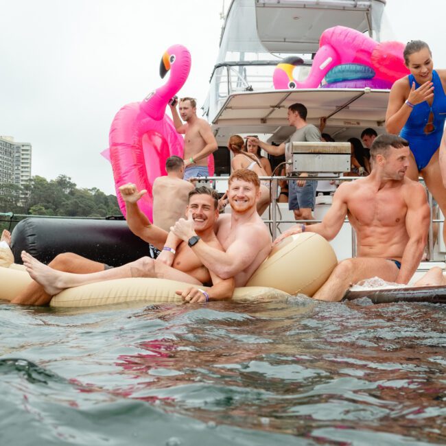 A group of people enjoying a party on a yacht adorned with inflatable flamingos. Two individuals in the foreground are lounging on large inflatable pool toys in the water, smiling and posing for the camera. Other party-goers are interacting and dancing energetically in the background.