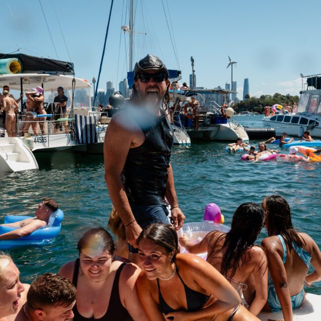 A group of people enjoy a sunny day on the water, some on boats and others on floating devices, with a city skyline and wind turbines in the background. Among them, a private yacht charter Sydney Harbour adds a touch of elegance to the scene.