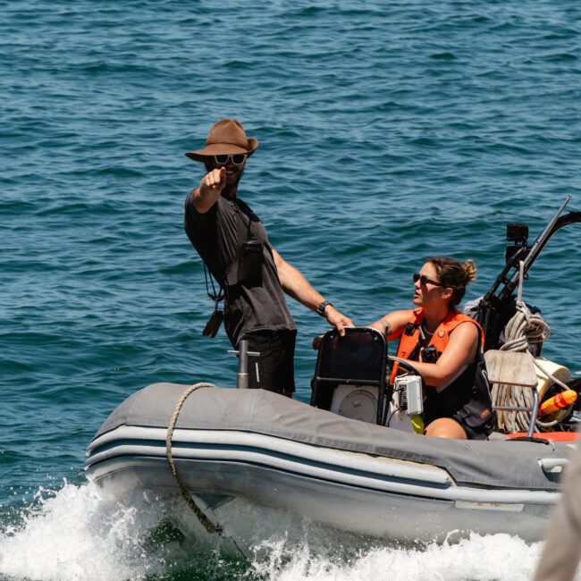 Two people are in an inflatable boat on a sunny day. One, wearing a brown hat, stands and points toward something in the distance. The other, donning sunglasses and a life vest, is seated at the controls of the boat. The peaceful water gleams under the bright sunshine.