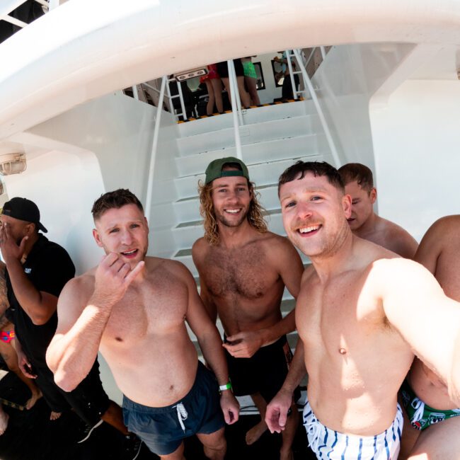 A group of shirtless men are posing and smiling for a selfie on a boat during a sunny day. Some are looking at the camera while others engage in conversation. In the background, there's a staircase and other people enjoying the scene.