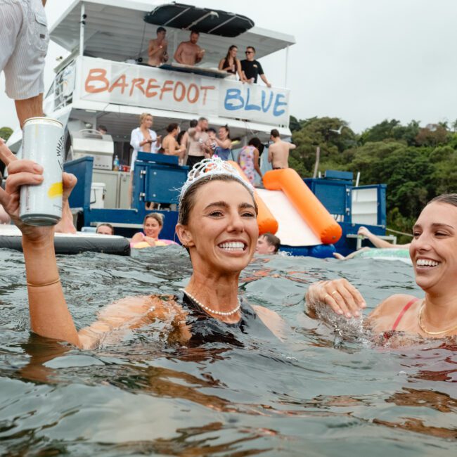 Two women smiling and standing waist-deep in water near a party boat labeled "Barefoot Blue." One woman, wearing a tiara and holding a drink, is laughing. Groups of people are visible on the boat and floating in the water, enjoying the festive ambiance under sunny skies.