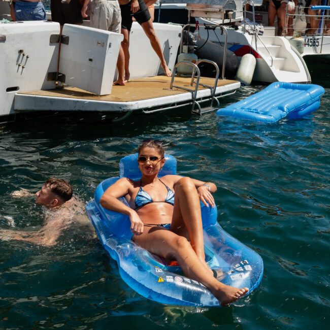 A woman in a bikini lounges on a blue inflatable float in the water near a boat, surrounded by other people swimming and standing on the boat, enjoying their luxury yacht hire Sydney adventure.