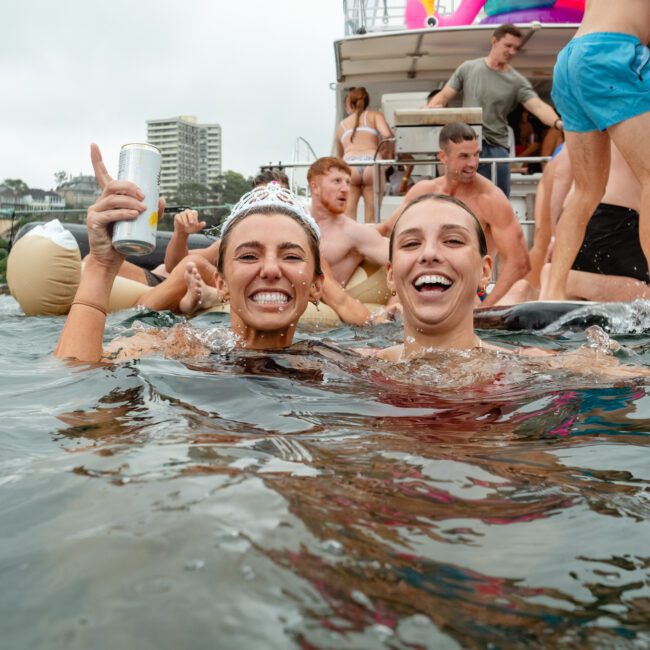 Two women are smiling and holding cans of White Claw hard seltzer while standing in water. A group of people on a boat and inner tubes are in the background, enjoying a lively atmosphere. Some buildings and trees are visible in the distance, enhancing the scenic view.