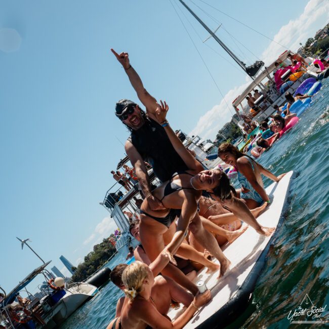 A group of people in swimsuits are on a paddleboard in the water, having fun. Some stand with arms raised, others lie down. Boats and a crowded dock form the background under a clear, sunny sky—perfect for corporate boat events Sydney or luxury yacht hire Sydney.