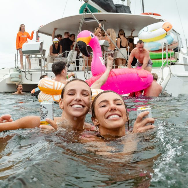 Two smiling women in a body of water holding drinks with a boat and people on inflatables in the background. The scene is lively with various pool toys on a catamaran, including a large pink flamingo float. The atmosphere is festive and joyful, perfect for making holiday memories.