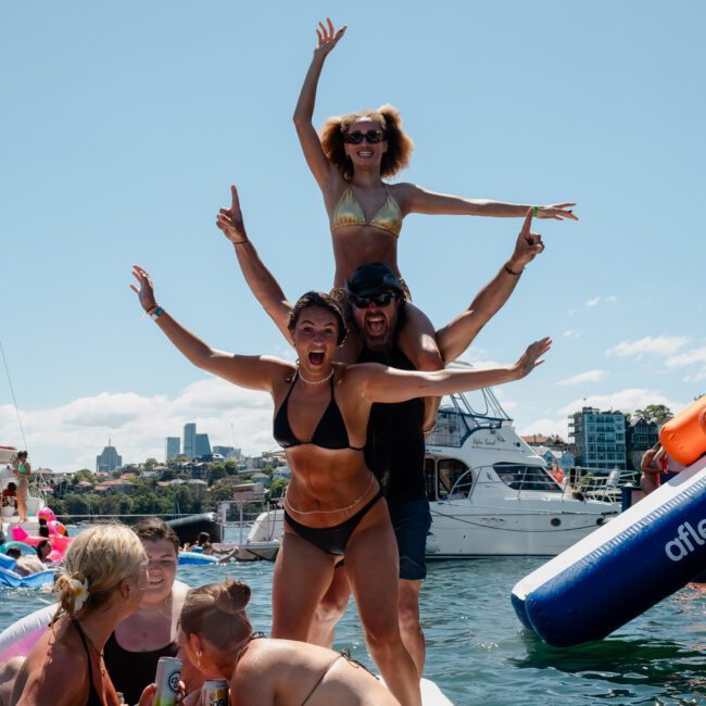 Four people form a human pyramid on a floating platform in the water, with luxury yachts and catamaran parties visible around them. Other boats and lively crowds enhance the vibrant atmosphere.