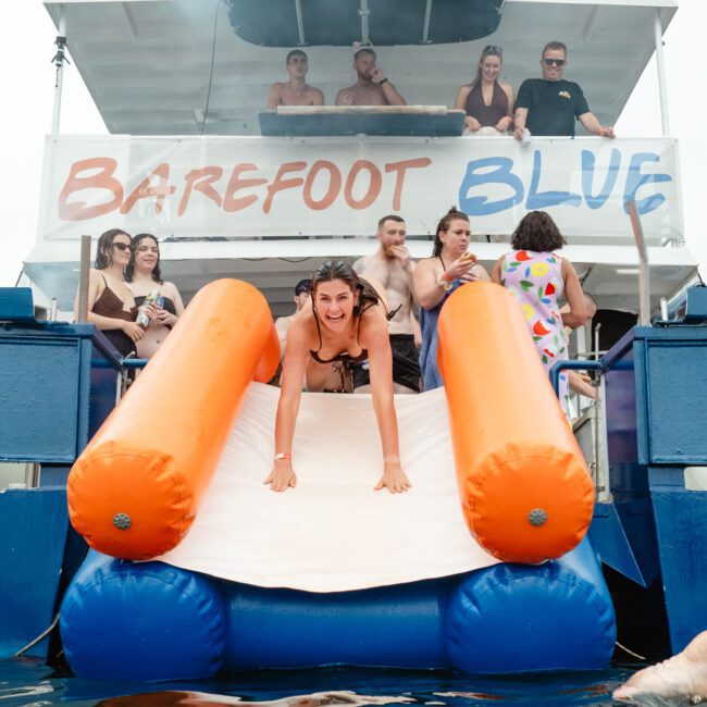 A woman joyfully slides down a water slide from a boat named "Barefoot Blue." Several people are standing and sitting on the boat, watching and enjoying the scene. The atmosphere is casual and fun, suggesting a day of leisurely water adventures.