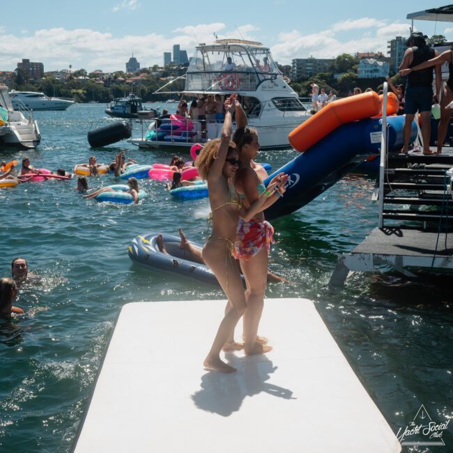 Two women dance on a dock next to a floating water slide, while people swim and lounge on inflatables in the water, with more guests enjoying themselves on boats in the background. It looks like an ideal setting for a Sydney boat party hire!