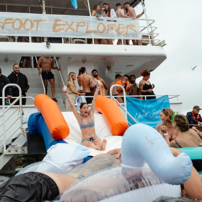 A group of people wearing swimsuits are having fun on a boat named "Barefoot Explorer." Some people are on the boat's upper deck, while one person slides down an inflatable water slide into the water. Others are swimming around and lounging on colorful inflatables.