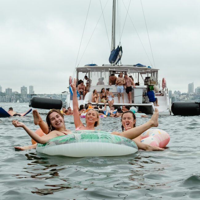 A group of people are enjoying a day out on the water, with many floating on inflatable tubes and swimming. Three people are sitting together on an inflatable tube, smiling and waving. In the background are moored boats and a picturesque city skyline reflecting in the water.