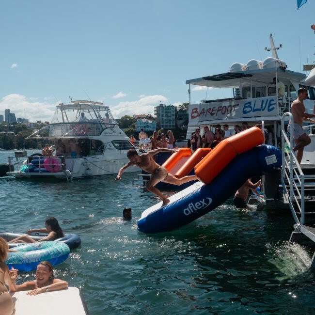 People are enjoying various water activities from a boat named "Barefoot Blue" on a sunny day, with some jumping off inflatables and others swimming or lounging on floats in the water. It feels like the perfect Sydney boat party hire.